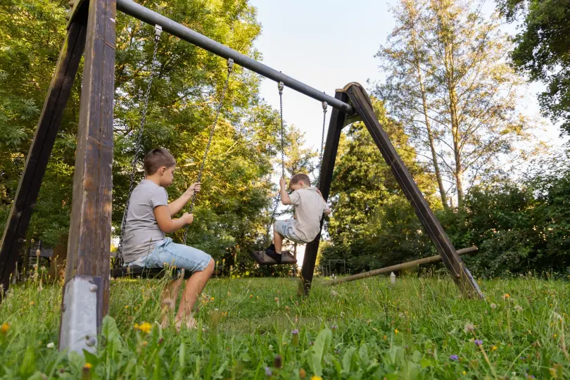 Two children play on wooden swings in a grassy outdoor area surrounded by trees, enjoying the well-maintained landscape on a sunny day.