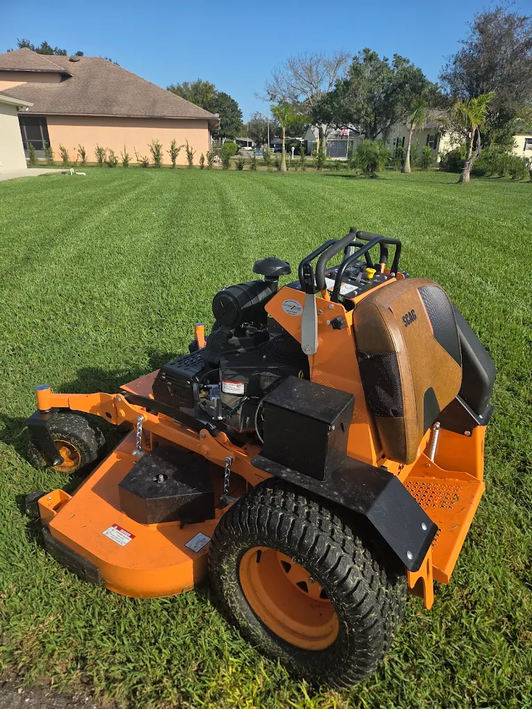 An orange stand-on commercial lawn mower, perfect for professional lawn care, is parked on a neatly mowed grassy lawn, with houses and trees enhancing the attractive landscape in the background.
