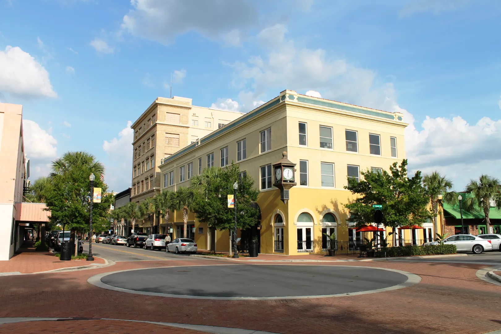A yellow three-story building stands at a street corner with a small roundabout, clock tower, well-kept landscape, parked cars, and trees under a partly cloudy sky.
