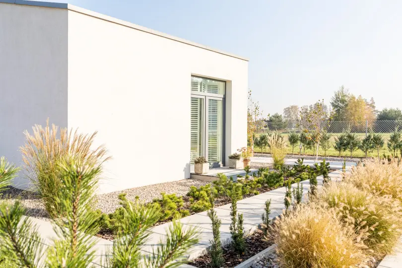 A modern, white, flat-roofed house with large glass doors is surrounded by expert landscape design featuring manicured shrubs, grasses, and a gravel pathway on a sunny day.