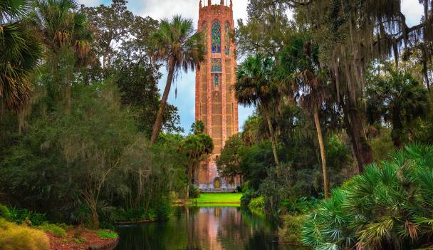 A tall, ornate stone tower stands behind a pond, nestled within a beautifully maintained landscape of dense green trees and tropical plants.
